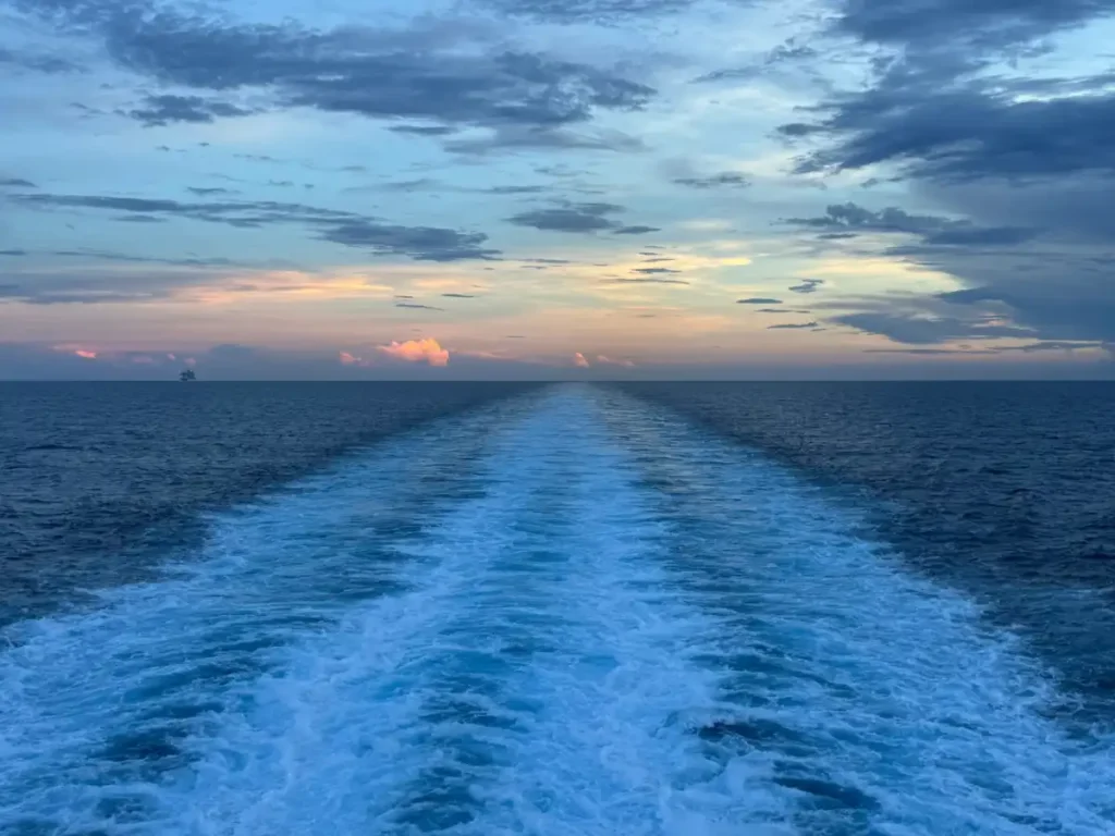The churning water wake trailing behind the back of a moving cruise ship at dusk.